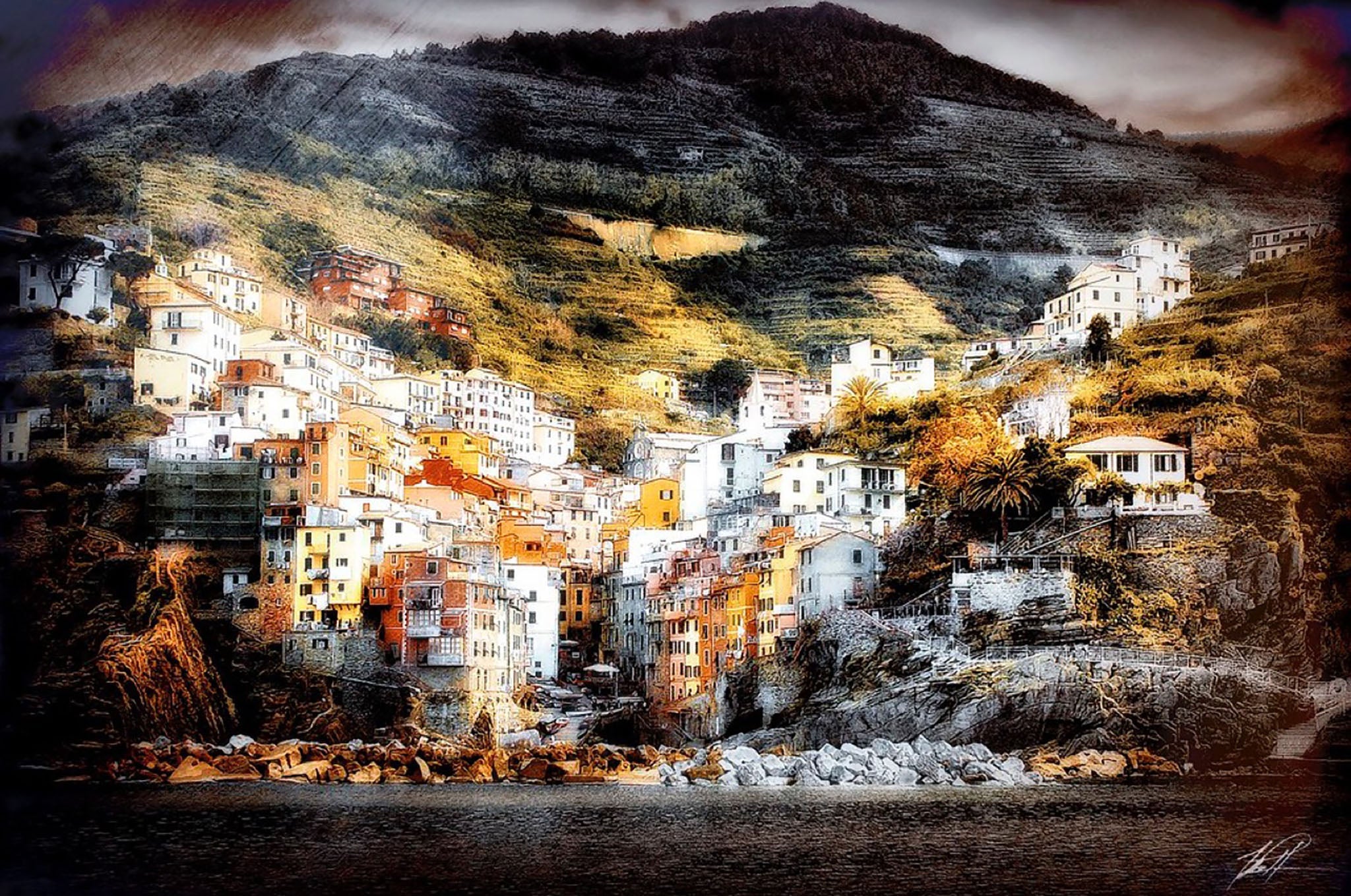 A photograph depicting a town in the bottom of a deep valley next to a body of water. The town is made up of multiple colorful buildings that are a wonderful contrast to the darkness of the hill behind them. There is a filter over the photograph causing the image to appear grainy. 18"x12" photography by Tom Patterson